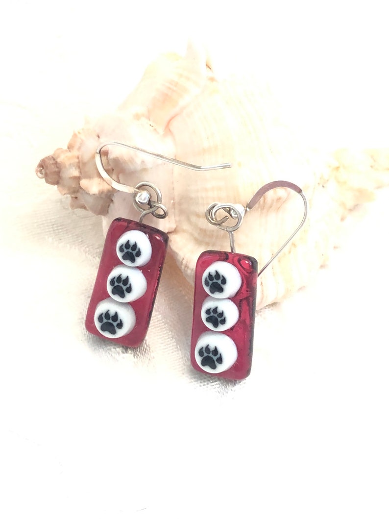 May include: Red glass dangle earrings with silver hooks and three white circles with black paw prints on each earring.