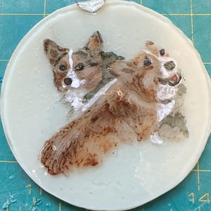 May include: A hand-painted glass ornament featuring two brown and white corgi dogs. The dogs are facing different directions, with one looking directly at the viewer. The ornament is round and has a white background.