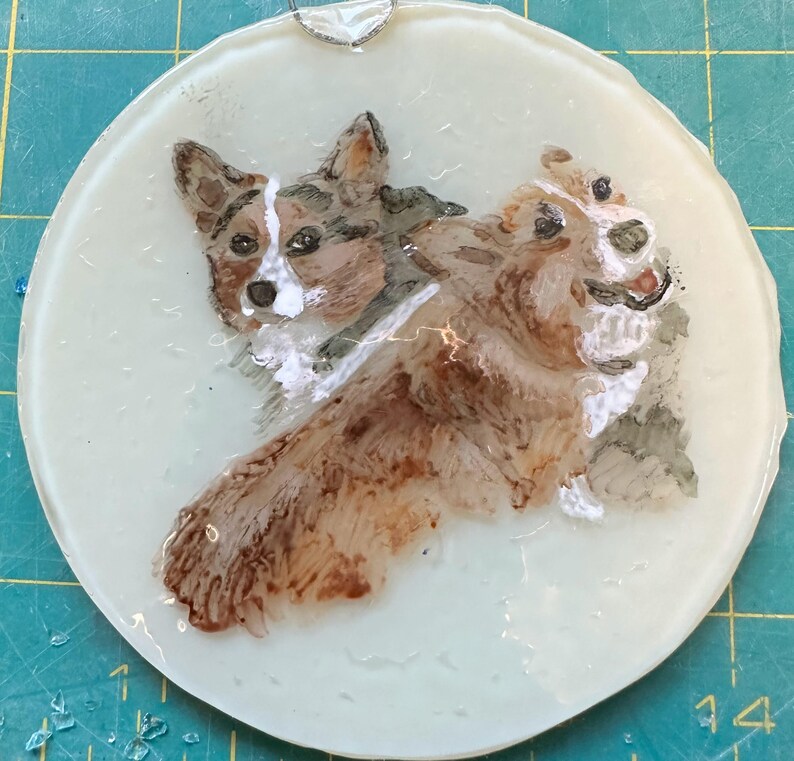 May include: A hand-painted glass ornament featuring two brown and white corgi dogs. The dogs are facing different directions, with one looking directly at the viewer. The ornament is round and has a white background.