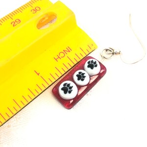 May include: Red glass earrings with three white paw print charms. The earrings are approximately 1 inch long and have silver hooks.