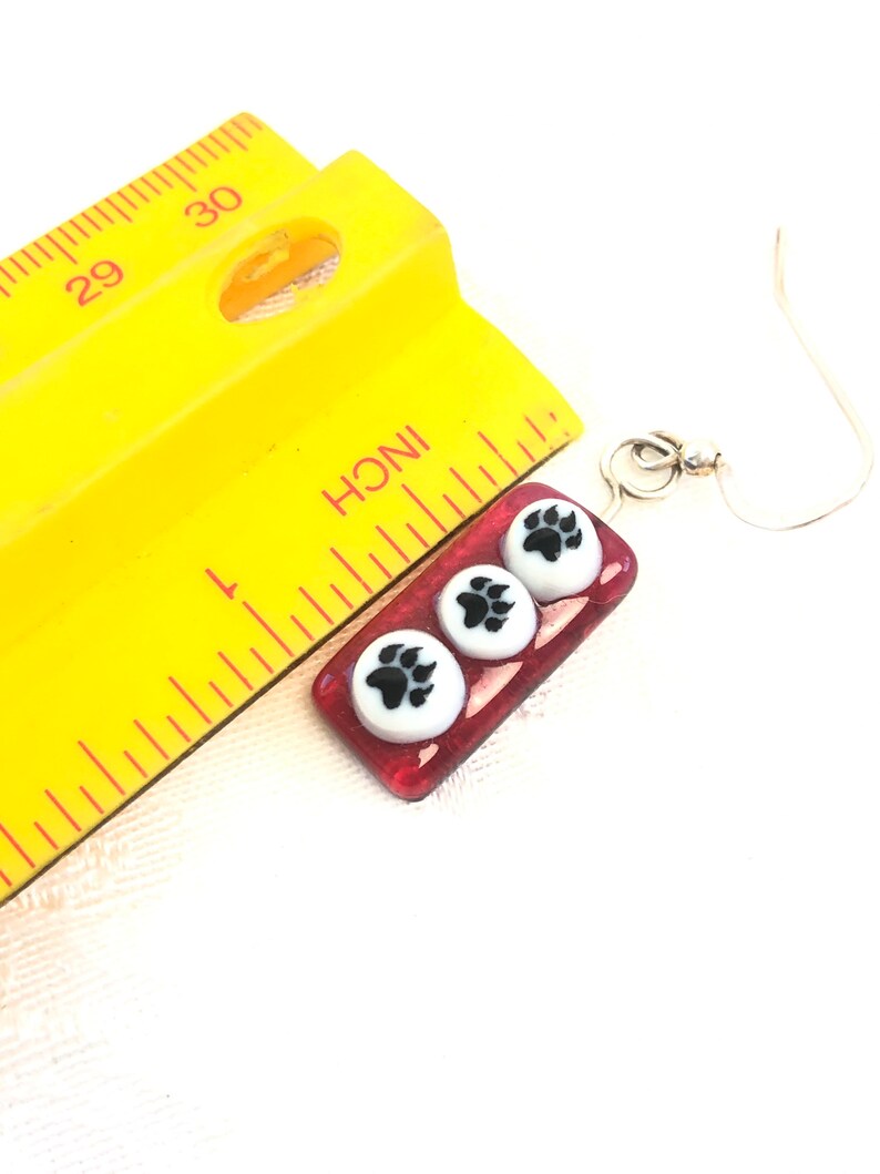 May include: Red glass earrings with three white paw print charms. The earrings are approximately 1 inch long and have silver hooks.