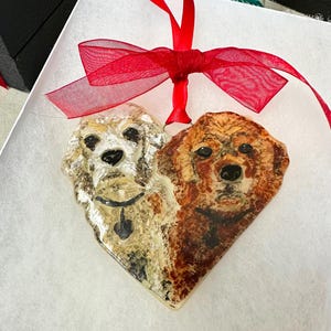 May include: A heart-shaped glass ornament with a painted image of two dogs, one white and one brown, with their heads facing forward. The ornament is tied with a red ribbon.