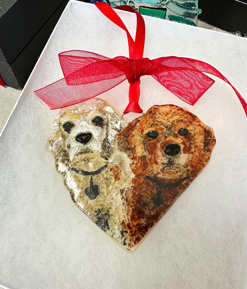 May include: A heart-shaped glass ornament with a painted image of two dogs, one white and one brown, with their heads facing forward. The ornament is tied with a red ribbon.