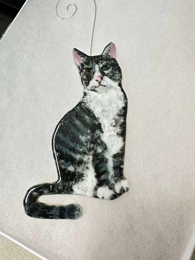 May include: A glass ornament of a black and white cat with green eyes. The cat is sitting with its tail curled around its legs. The ornament has a silver hanger.