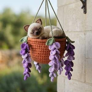 Könnte beinhalten: Eine gehäkelte Katzenfigur ruht in einem hängenden Korb, der mit lila und grünen floralen Akzenten verziert ist. Der Korb wird von grünen Schnüren an einer schwarzen Metallhalterung befestigt, die an einer Steinmauer befestigt ist. Die Katze ist beige und braun.