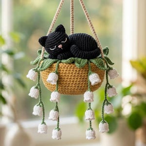 Puede incluir: Una maceta colgante de ganchillo con un gato negro durmiendo. La maceta es de color amarillo mostaza con hojas verdes y detalles de flores blancas. El gato está acurrucado en la maceta, suspendido por una cuerda beige.