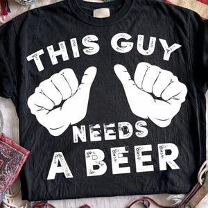 May include: A black short-sleeved t-shirt featuring a white graphic of two hands with thumbs pointing inward. The text printed in white reads THIS GUY NEEDS A BEER, with the words positioned above and below the illustrated hand graphics.