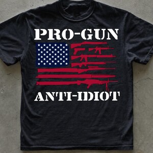 May include: A black short-sleeved t-shirt featuring a graphic of the American flag where the stripes are replaced by various firearm silhouettes. The text PRO-GUN is printed in white above the flag, and ANTI-IDIOT is printed in white below the flag.