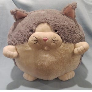 May include: A plush, round cat toy in shades of gray and cream. The cat has a pink nose, embroidered whiskers, and small, upright ears. The toy is designed with a soft, cuddly appearance, perfect for children or cat lovers.