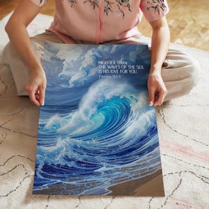 Könnte beinhalten: Ein lebendiger Druck mit einer dynamischen Meereswelle in Blau- und Weißtönen. Das Kunstwerk enthält den Text "MIGHTIER THAN THE WAVES OF THE SEA IS HIS LOVE FOR YOU. Psalm 93:4". Der Druck wird von einer Person gehalten.