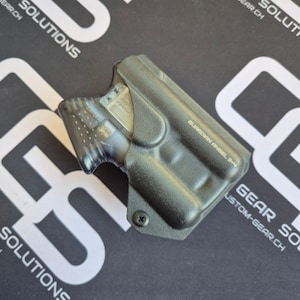 Könnte beinhalten: Ein schwarzes, geformtes Holster mit strukturierter Oberfläche und einer einzelnen schwarzen Schraube an der Basis. Das Holster ist mit dem Text GUARDIAN ANGEL 3+4 geprägt und enthält ein teilweise sichtbares, graues, durchscheinendes Gerät.