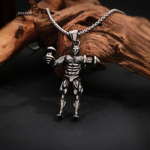 May include: Silver-toned pendant necklace featuring a muscular weightlifter holding dumbbells. The pendant is detailed with anatomical features and hangs from a chain. The background includes a piece of wood and a dark surface.