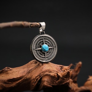 May include: A silver compass pendant featuring a turquoise stone at its centre. The compass is encircled by a circular design with Roman numerals and directional markings.