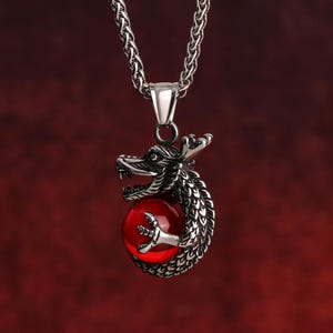 Peut inclure: Un collier en chaîne argent avec un pendentif représentant un dragon tenant une pierre précieuse rouge.
