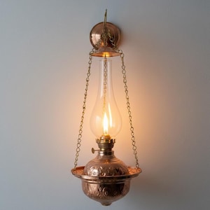 Może przedstawiać: Miedziana lampa naftowa ścienna z przezroczystym szklanym kloszem i zapalonym płomieniem. Lampa ma dekoracyjny, wytrawiony wzór na podstawie i jest zawieszona na łańcuchach. Ozdobne oświetlenie.