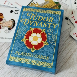 May include: A blue box of Tudor Dynasty playing cards with a red rose and gold accents. The box has the words "Tudor Dynasty" and "Playing Cards" printed on it.