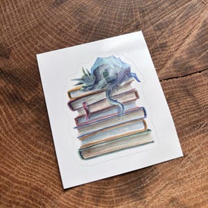 May include: A white sticker featuring a watercolor illustration of a blue dragon resting on a stack of books. The books are various colors, including brown, orange, and red. The dragon has a long tail and is curled up on top of the books.