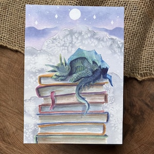 May include: Watercolor art print of a sleeping dragon atop a stack of books. The dragon is blue-green with a spiky back, resting on books with red and orange spines. The background features a moon, stars, and clouds in shades of purple and white.