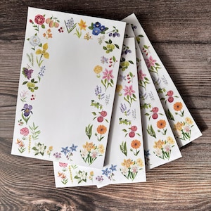 May include: A set of four floral stationery note cards with a white background and a colorful floral border. The border features a variety of flowers, leaves, and berries in shades of pink, red, yellow, blue, and green.
