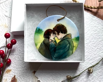 May include: A round ceramic ornament with a painted image of a couple embracing. The couple is depicted in a romantic pose, with the woman in a brown dress and the man in a blue coat. The background is a watercolor wash of blue and green, suggesting a landscape.