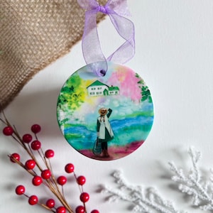 May include: A round ceramic ornament with a watercolor painting of a woman in a straw hat and a green dress standing by a lake. The background features a green house and colorful trees.