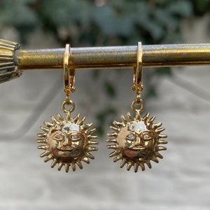 Op de afbeelding: Een paar gouden oorbellen met een zonontwerp met een gezicht. De oorbellen hangen aan een gouden staaf.
