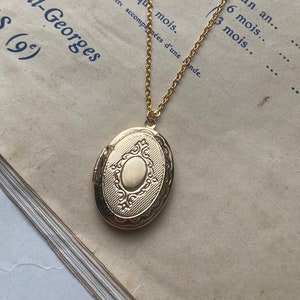 May include: A gold locket necklace with an oval-shaped locket. The locket has a detailed design with a smaller oval in the centre. The necklace is lying on a vintage book with text about subscriptions.