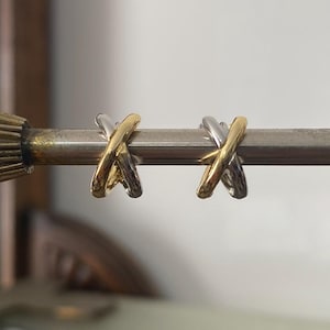 May include: A pair of gold and silver-toned X-shaped earrings. The earrings are displayed on a metal rod against a neutral background. The earrings have a polished finish and a modern design.