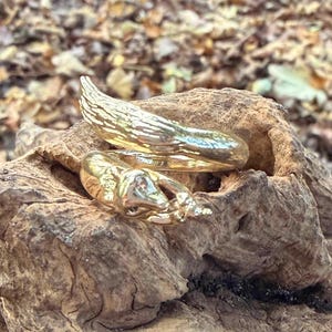 May include: Two gold-colored rings rest on a piece of weathered wood. One ring features a textured, wing-like design, while the other has a detailed, sculpted appearance. The rings are set against a backdrop of fallen leaves and a blurred forest.