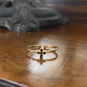 Può includere: Un anello d'oro con un piccolo design a croce nera. L'anello è su una superficie di legno.