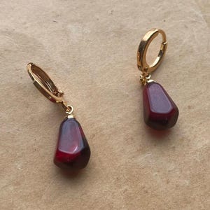 Puede incluir: Un par de pendientes de aro dorados con un colgante de piedra en forma de lágrima de color rojo oscuro. Los pendientes se muestran sobre un fondo de papel vintage con texto y un emblema decorativo.