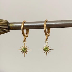 Könnte beinhalten: Goldene Creolen mit einem baumelnden Stern-Anhänger, der einen grünen Edelstein in der Mitte trägt.