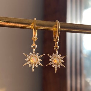 May include: A pair of gold hoop earrings with a dangling star charm. Each star has a clear opal centre and is surrounded by smaller clear stones.