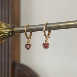 Peut inclure: Une paire de créoles dorées avec un pendentif en forme de cœur rouge. Les boucles d'oreilles sont suspendues à un présentoir en métal.