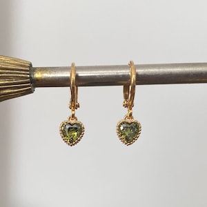 K&ouml;nnte beinhalten: Ein Paar goldene Creolen mit kleinen herzf&ouml;rmigen Anh&auml;ngern, die mit olivgr&uuml;nen facettierten Steinen besetzt sind. Jeder Herzanh&auml;nger ist von einem zarten, perlenartigen Goldrand umgeben und h&auml;ngt an einem schlichten goldenen Ohrringverschluss.