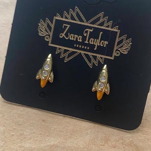 May include: Gold-coloured rocket earrings with orange detailing and clear gems, presented on a black Zara Taylor branded card. The card is set against a vintage paper background with musical notes and text.