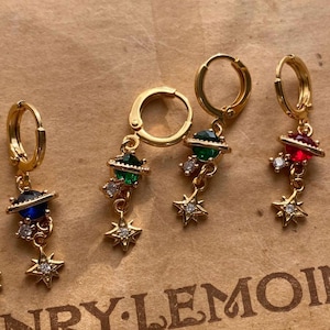 May include: Five pairs of gold hoop earrings with dangling charms. Each charm features a small, colorful planet with a star-shaped design. The planets are blue, green, red, and blue.