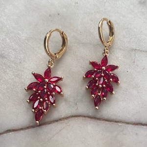 Könnte beinhalten: Ein Paar goldene Creolen mit herabhängenden roten Edelstein-Clustern. Die Edelsteine sind in Tropfenform angeordnet.