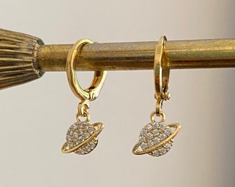 May include: A pair of gold hoop earrings with a dangling charm of a small, sparkly planet. The earrings are hanging on a gold display stand.