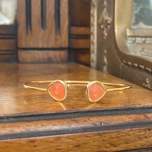 Peut inclure: Un bracelet jonc en or avec deux pierres en forme de larme de couleur corail. Le bracelet est posé sur une surface en bois.