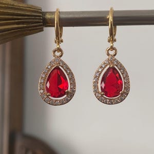 May include: A pair of gold-coloured hoop earrings featuring teardrop-shaped red gemstones. Each earring is encircled by a halo of small, clear crystals. The earrings are suspended from a metal rod.