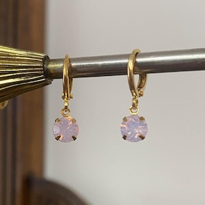 May include: Gold-coloured hoop earrings, each with a light purple, round gemstone. The earrings are displayed on a silver rod against a blurred background, creating a vintage aesthetic. The gemstones have a subtle, iridescent quality.