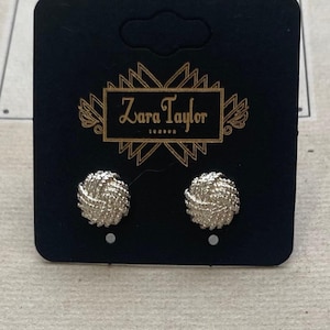 May include: A pair of silver-toned stud earrings with a textured, knotted circular design. The earrings are mounted on a black rectangular display card featuring the gold-colored text Zara Taylor London centered within a geometric frame.