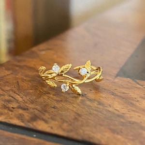 May include: A gold ring featuring a delicate vine design with small clear stones. The ring is resting on a wooden surface.