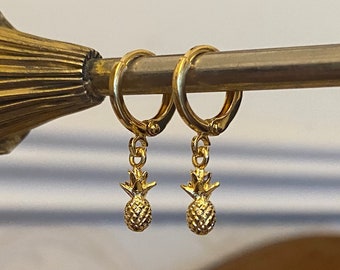 以下が含まれることがあります： 金色のフープピアス。それぞれのフープから小さなパイナップルのチャームがぶら下がっている。