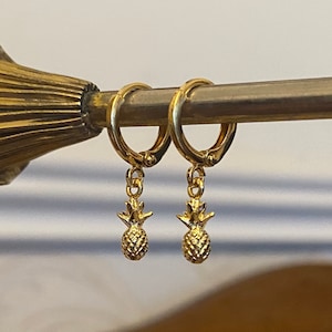 Könnte beinhalten: Ein Paar goldene Creolen mit einem kleinen Ananas-Anhänger, der an jeder Creole baumelt.