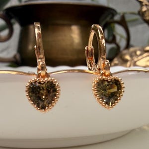 K&ouml;nnte beinhalten: Ein Paar goldfarbene Creolen mit herzf&ouml;rmigen Anh&auml;ngern, die facettierte olivgr&uuml;ne Steine enthalten. Jeder Herzanh&auml;nger ist von einem zarten, perlenartigen Goldrand umgeben und an einem kleinen Klappmechanismus f&uuml;r den Verschluss befestigt.