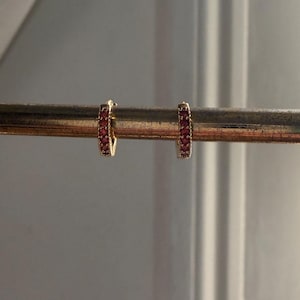 Peut inclure: Deux boucles d'oreilles créoles dorées, chacune incrustée de petites pierres rouges. Les boucles d'oreilles sont présentées sur une barre dorée, sur un fond neutre et doux, la lumière naturelle mettant en valeur les bijoux.