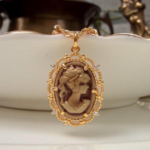 May include: A gold-toned pendant necklace featuring an oval-shaped cameo with a profile portrait of a person. The cameo is set within an ornate, filigree-style gold frame accented with small, clear, sparkling stones at the cardinal points.