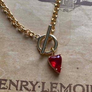 Può includere: Collana dorata con un ciondolo rosso a forma di cuore. La collana presenta una chiusura a levetta e un design a catena. Lo sfondo è una carta vintage con testo.